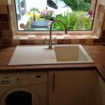 Kitchen sink with a view of the driveway through the window, above an integrated washing machine, nestled into the kitchen worktop.