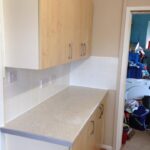 A newly installed kitchen countertop and cabinets with an unfinished wooden front, adjacent to a room with various items strewn about, showcases the expertise of a skilled kitchen fitter.