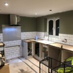 A kitchen under renovation with unfinished tile work and cabinetry, awaiting a kitchen fitter, and construction materials scattered around.
