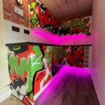 A vibrant graffiti-style mural decorates the walls of an urban kitchen space, enhanced with neon lighting accents.