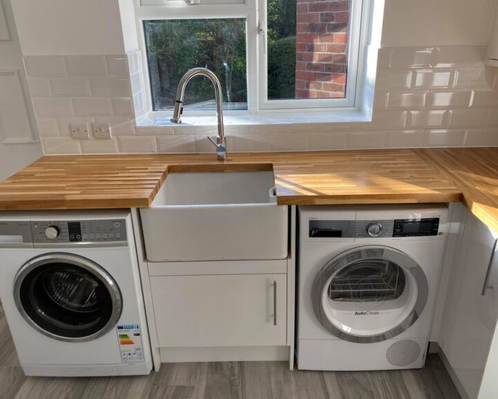A modern kitchen interior, showcasing a wooden kitchen worktop with an integrated white sink and a washing machine next to a dryer underneath.