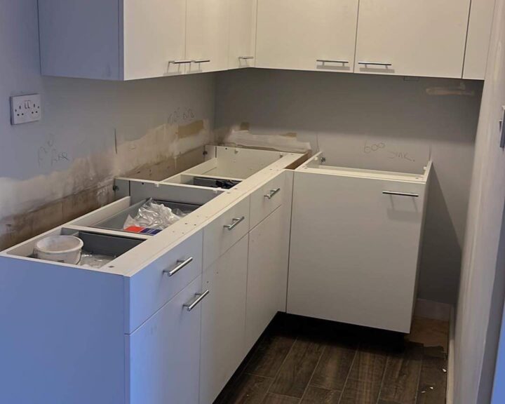 A modern kitchen in the midst of installation or renovation, featuring white cabinetry and a dark floor, with some construction debris and markings present. The kitchen worktop waits to be fitted into its designated space